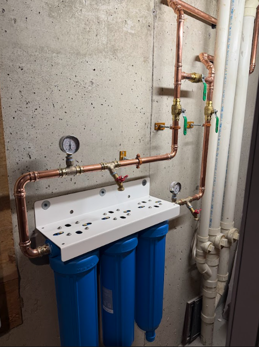 Wall-mounted water filtration system with three blue filter canisters and exposed copper pipes.