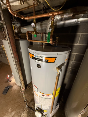 Water heater in a cramped utility closet with exposed pipes and a cinderblock wall