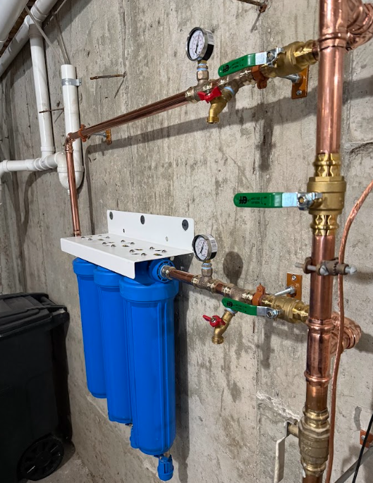 Basement wall with copper pipes, gauges, valves, and blue filter housing mounted on the left.