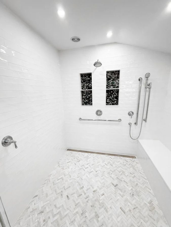 Bright white walk-in shower with tiled walls, herringbone floor, bench, and handheld showerhead.
