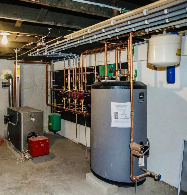 Basement utility room with boiler, pipes, water heater, and industrial equipment lining the wall