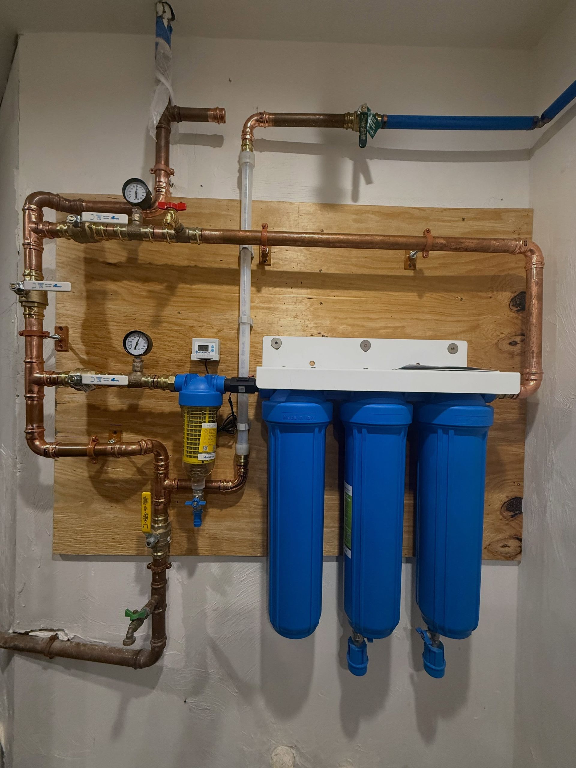 Wall-mounted water filtration system with blue filter cartridges, copper pipes, and gauges on a plywood board.