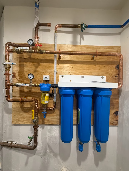 Wall-mounted water filtration system with gauges, valves, copper pipes, and three blue filter canisters.