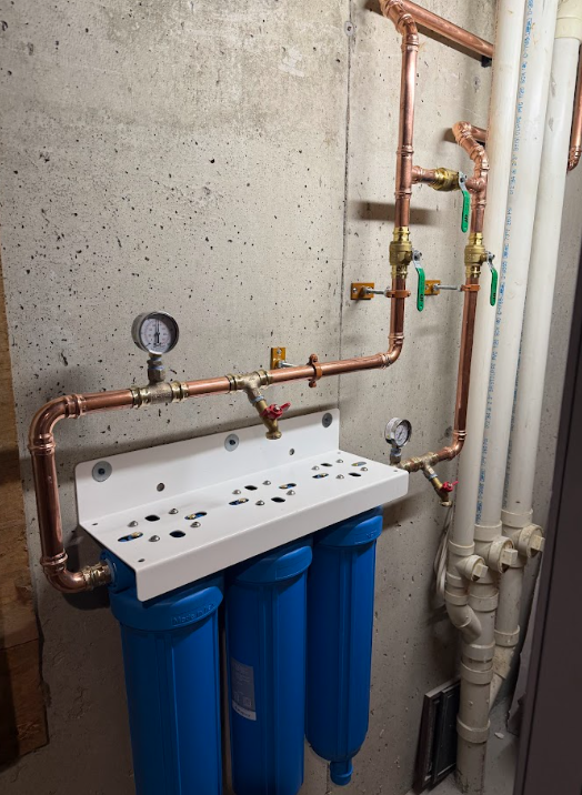 Water filtration system with copper pipes, pressure gauge, and blue filters mounted on a concrete wall.