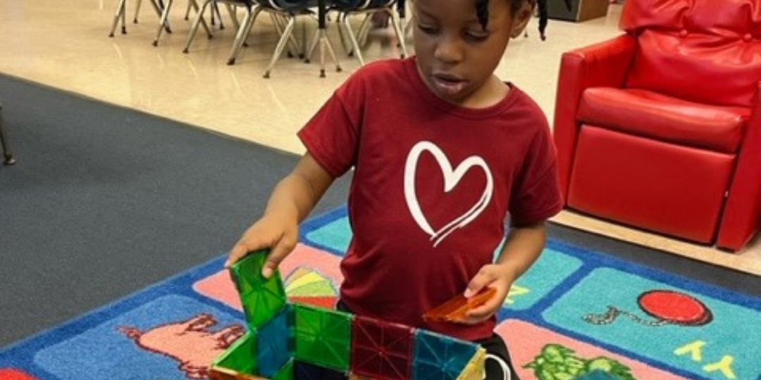 A little girl wearing a red shirt with a heart on it is playing with toys on the floor.