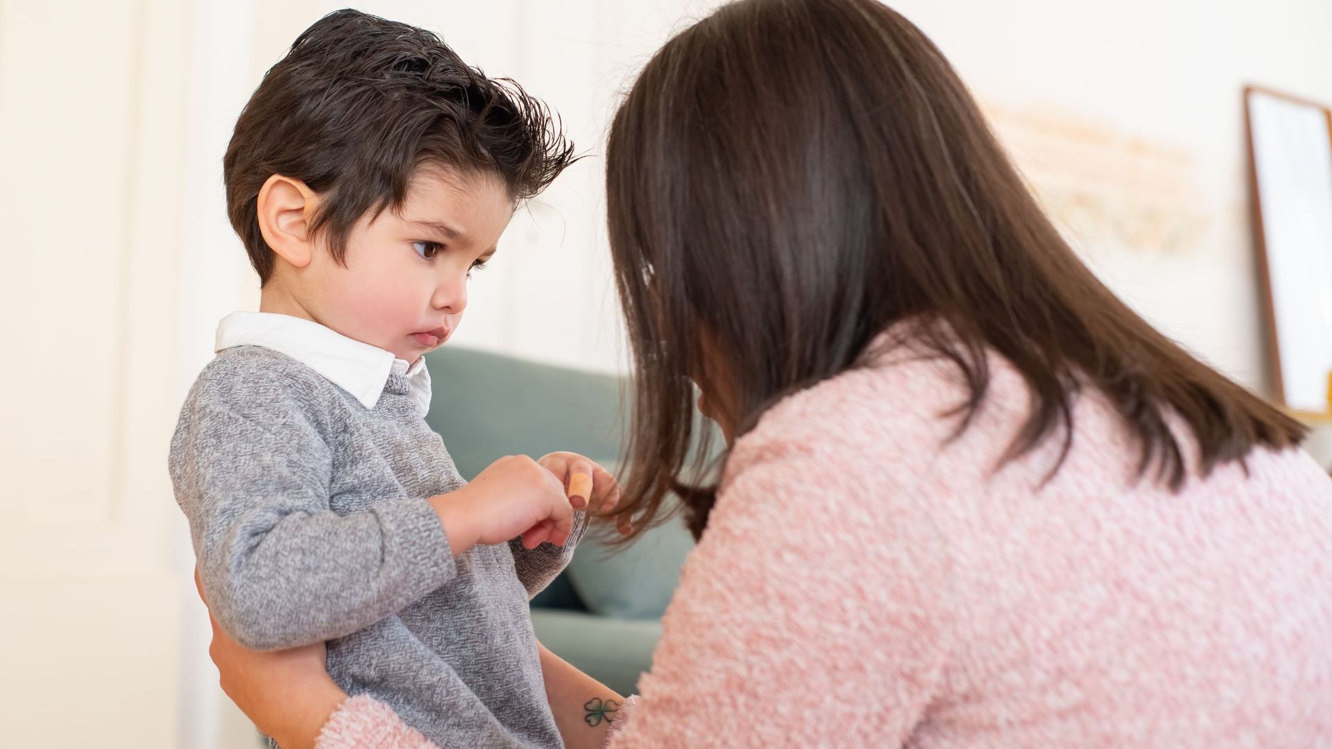 Woman holding a child; both looking down. The child wears a gray sweater. Warm-toned interior.