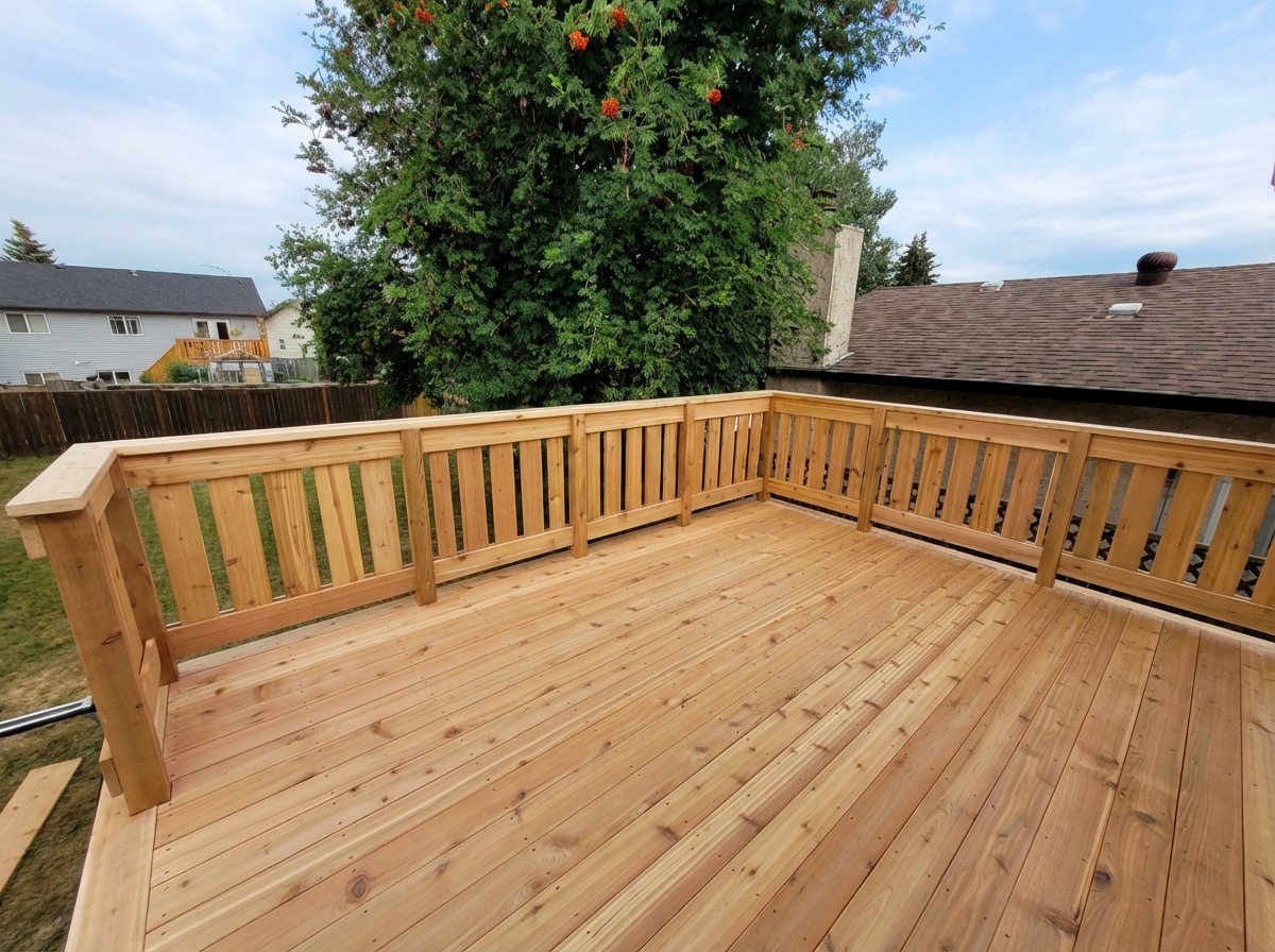 Wooden deck with railings overlooking a backyard, with a large tree and neighboring rooftops in view