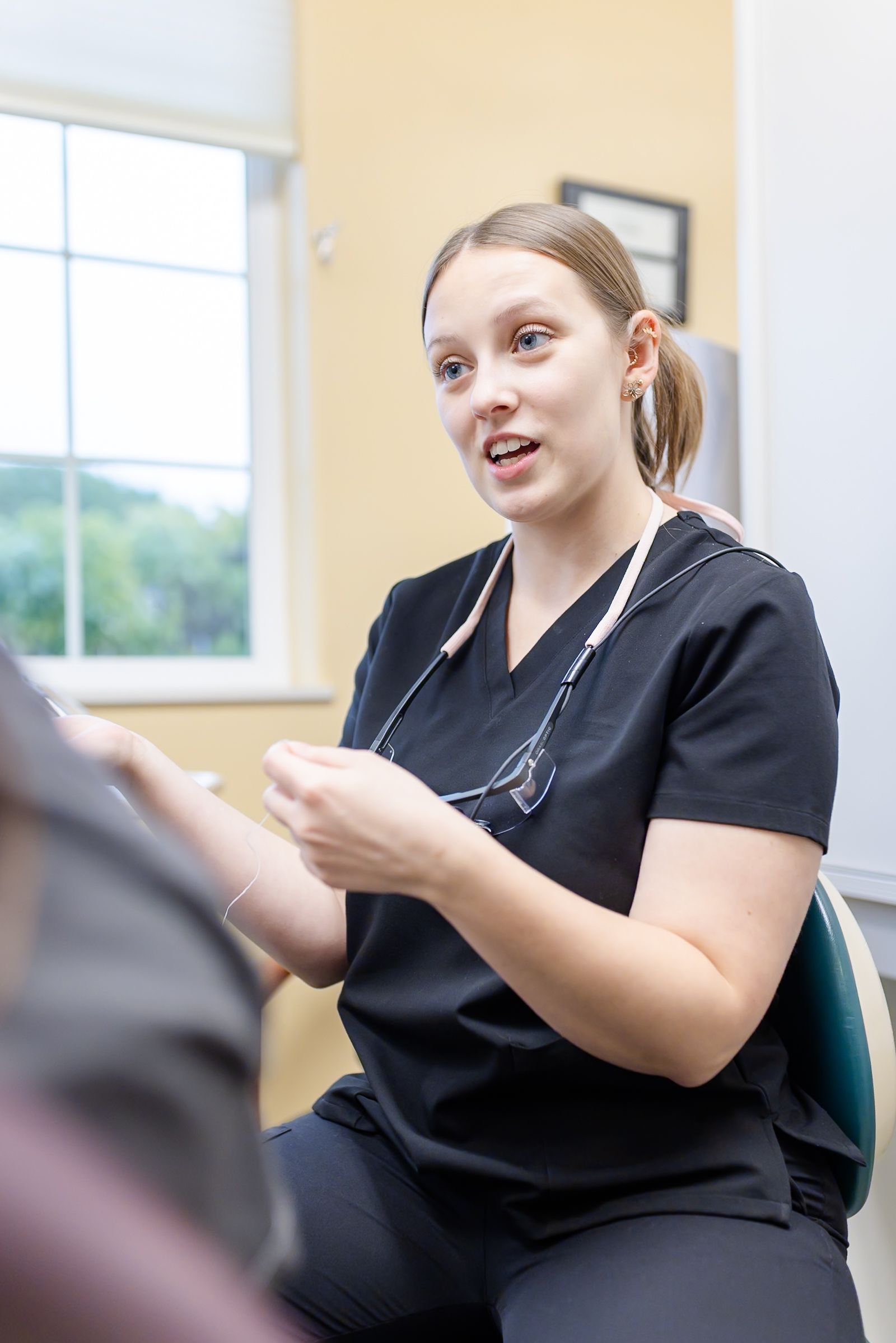 Dental hygienist in black scrubs gesturing, explaining dental floss to a patient in a light-filled office.