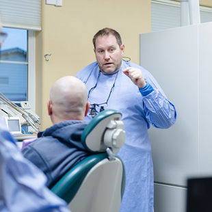Dentist in blue gown gesturing, explaining procedure to seated patient. Dental office setting.
