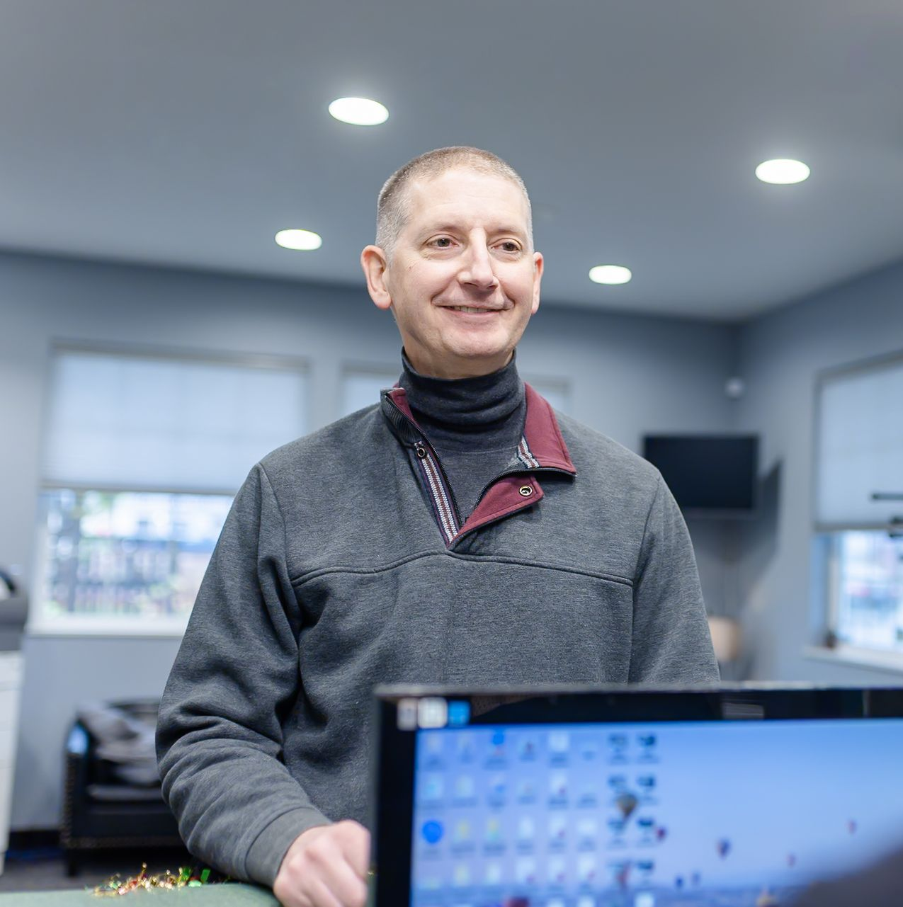 Man in gray sweater smiles, standing indoors near a desk with a computer screen.