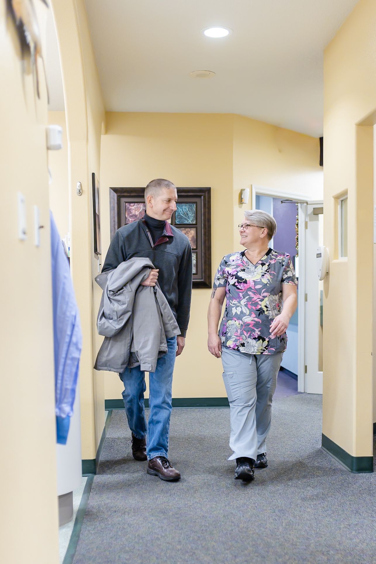 Man and woman walking down a hallway, smiling at each other. Woman wears scrubs, man holds a jacket.