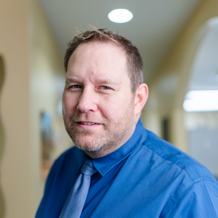 Man in blue shirt and tie smiling in a hallway.