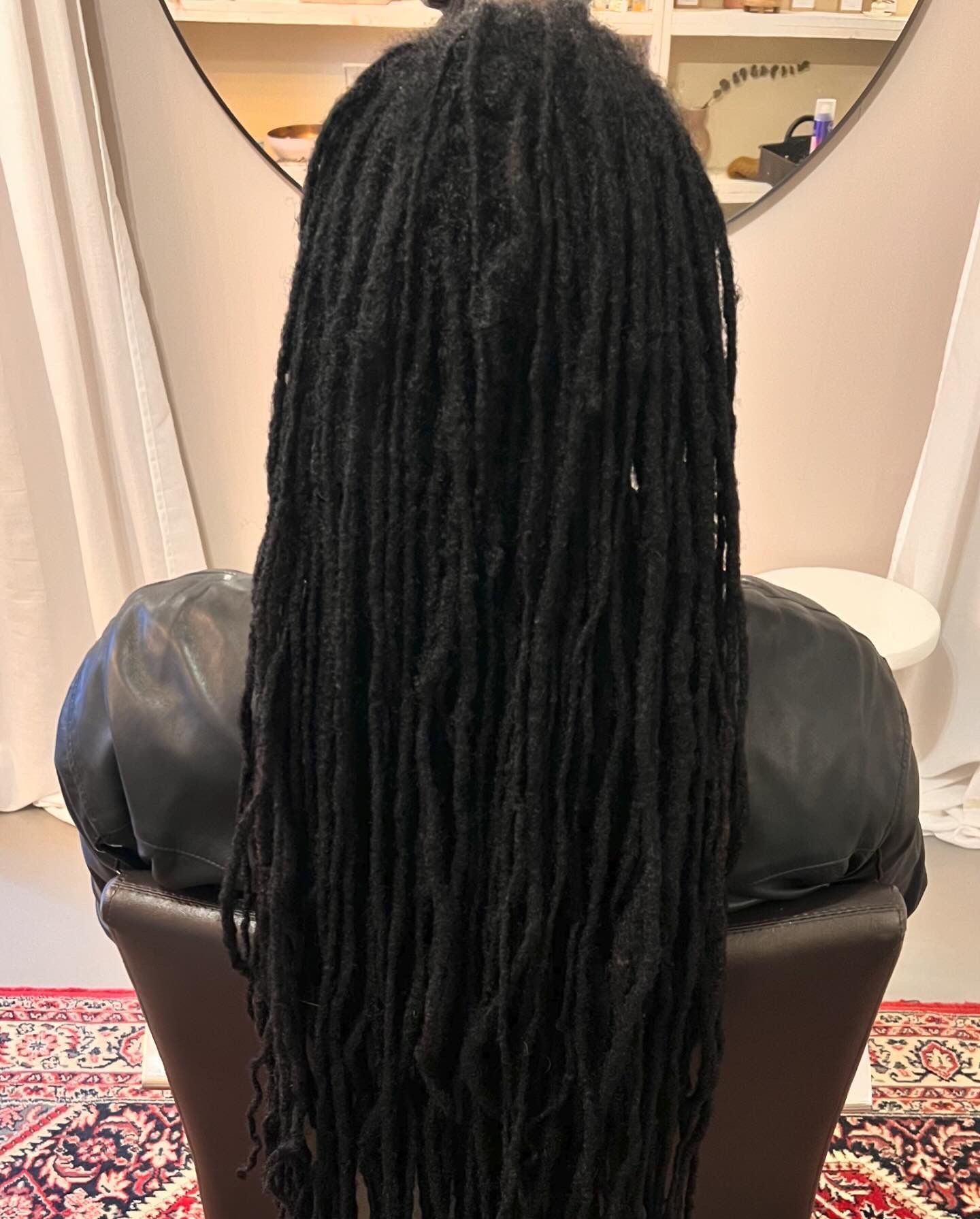 Een persoon met lange zwarte dreadlocks zit op een stoel, met een spiegel op de achtergrond.