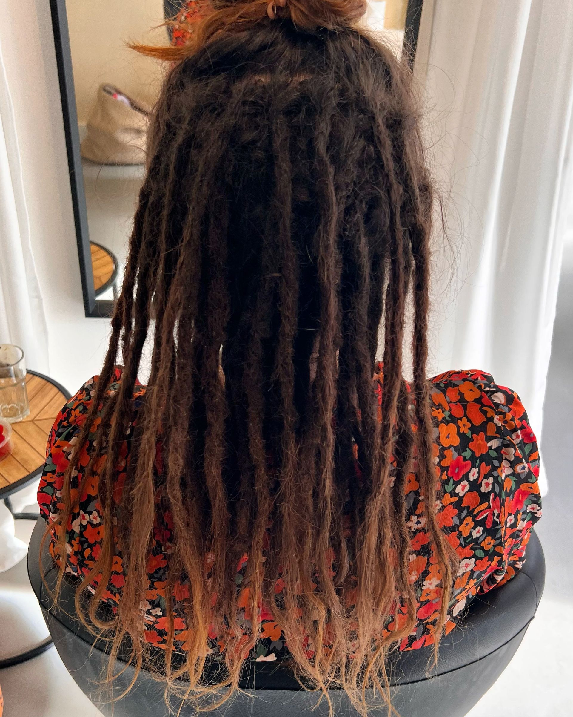Een persoon met donkerbruine dreadlocks, gekleed in een oranje bloemenblouse, zit op een stoel.