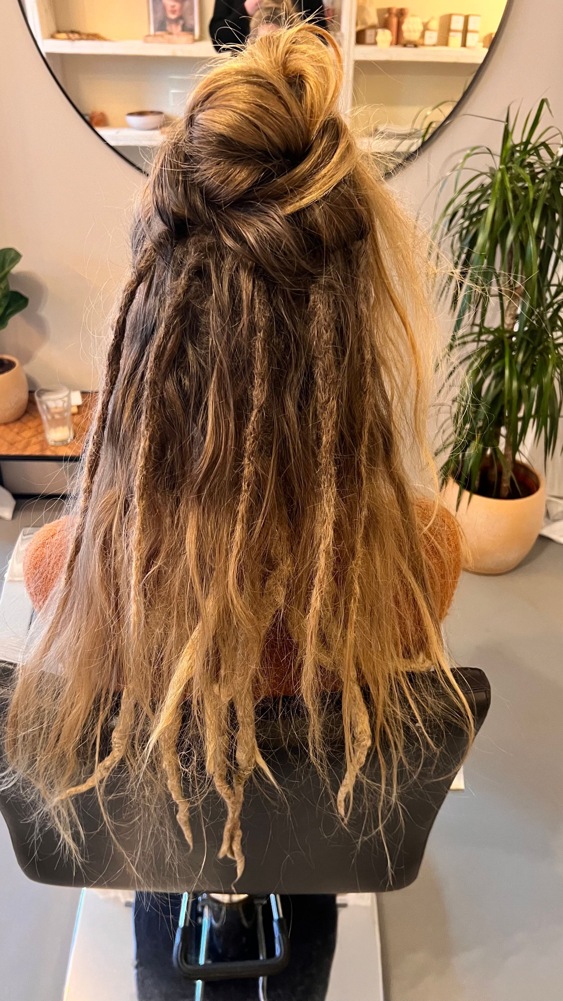 Vrouw met lang, gelaagd, gedeeltelijk gevlochten blond haar, met een knot bovenop, zit in een kappersstoel.