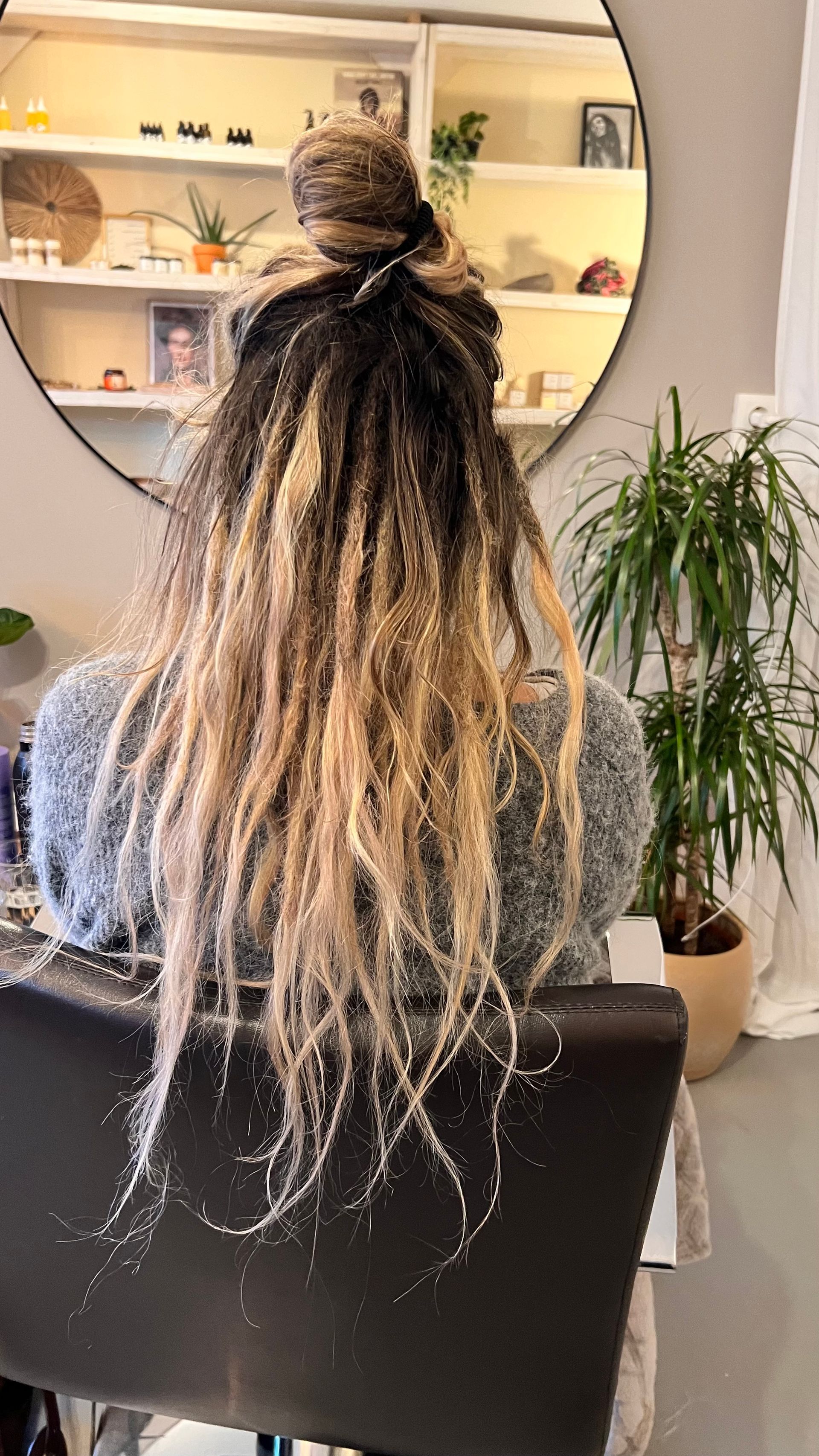 Persoon met lange, blonde en zwarte dreadlocks in een knot, zittend voor een spiegel, plant op de achtergrond.
