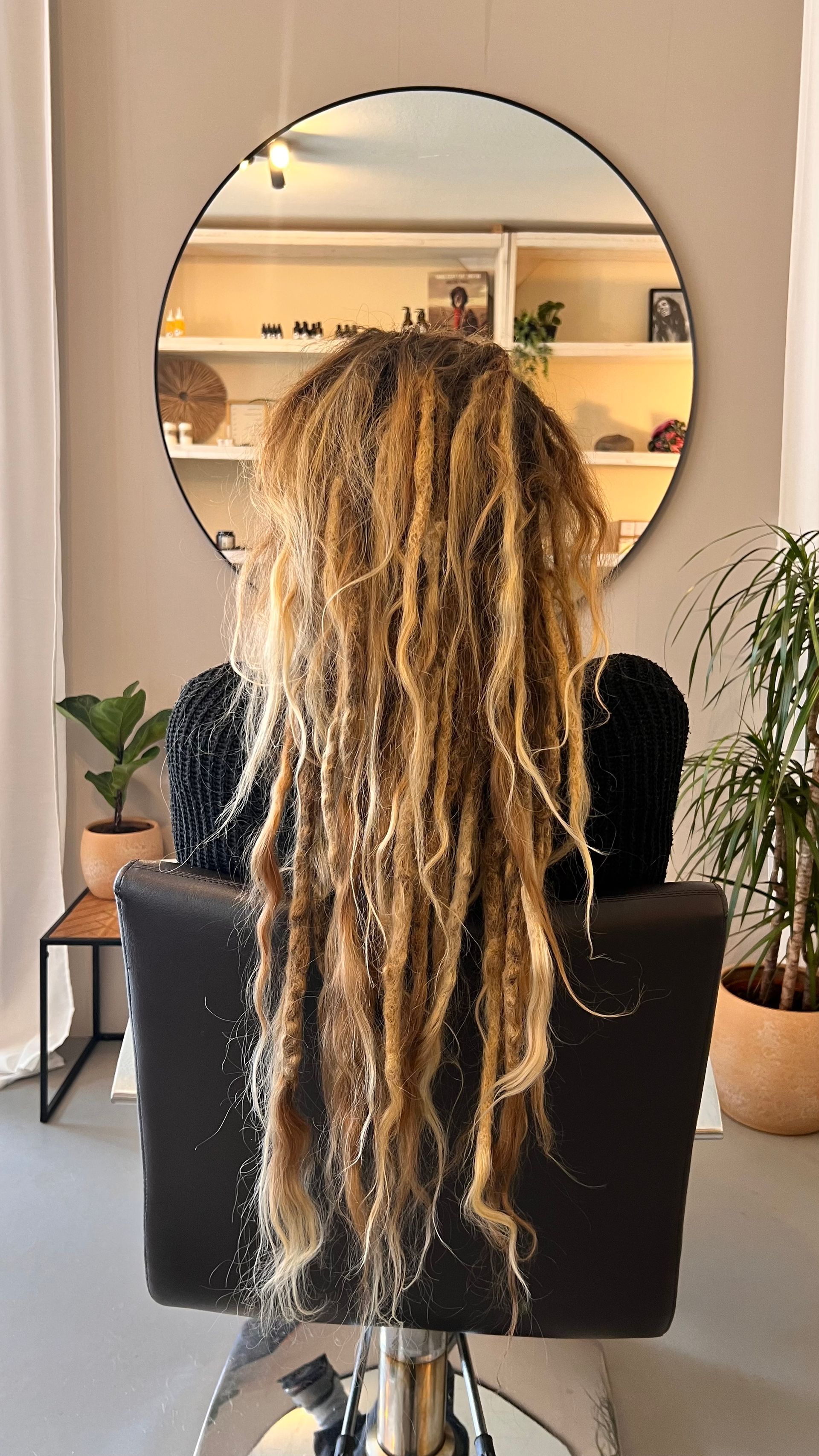 Een persoon met lange, blonde dreadlocks zit in een kappersstoel en kijkt weg van de camera.