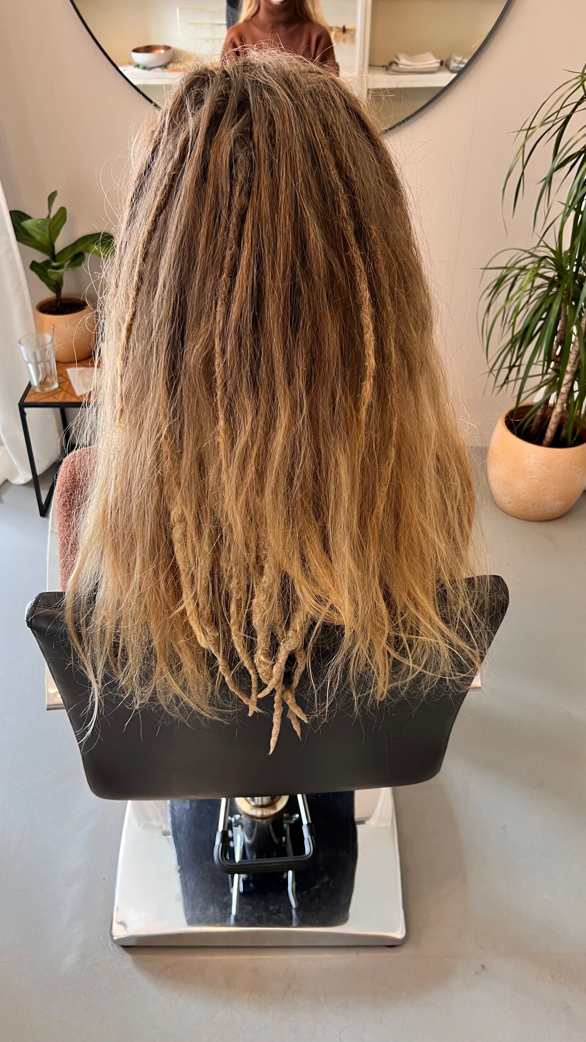 Een persoon met lang, golvend haar, dat van donkerbruin naar blond verandert, zit in een kappersstoel.