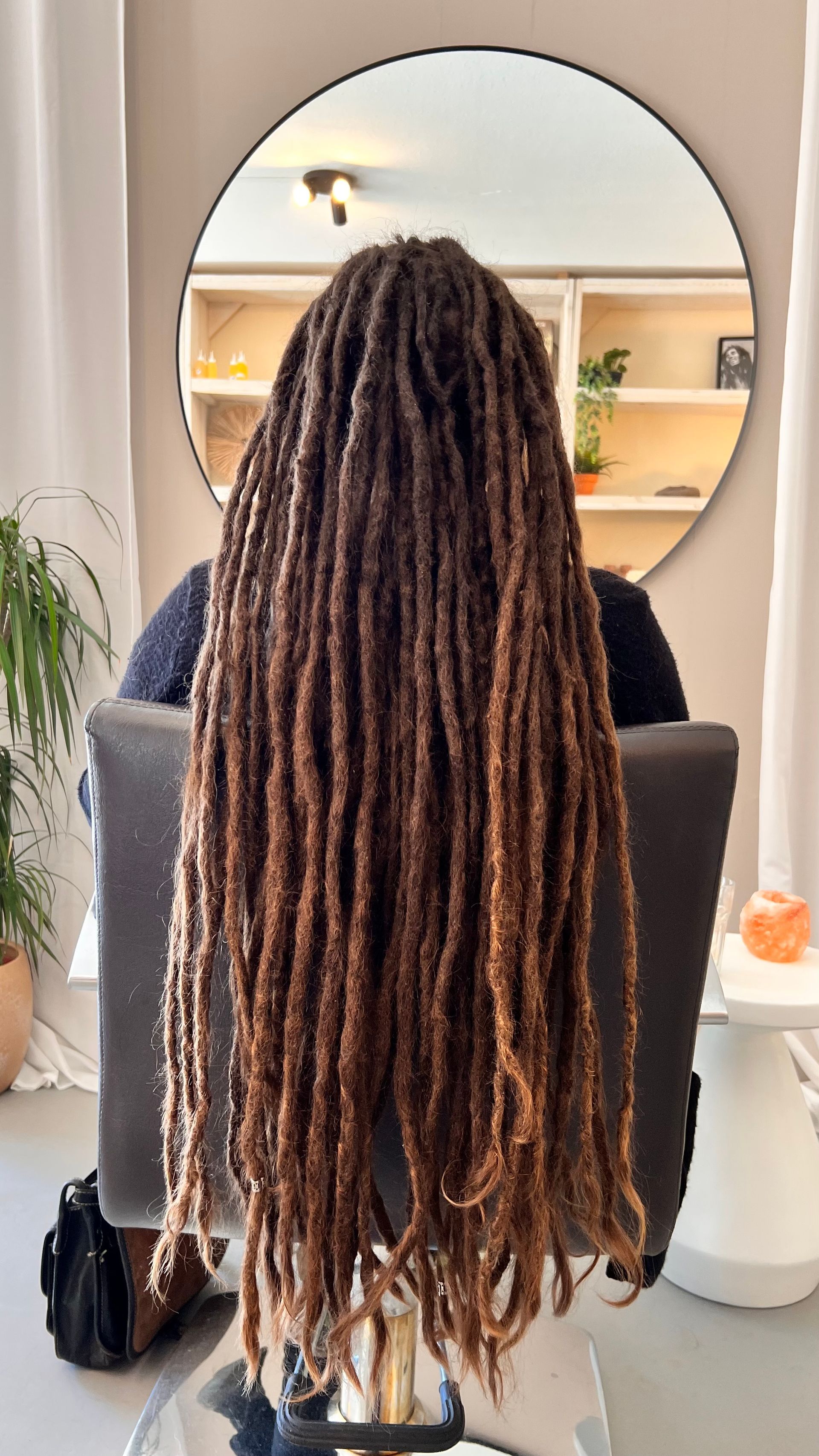 Een persoon met lange bruine dreadlocks zit op een stoel, met een ronde spiegel op de achtergrond.