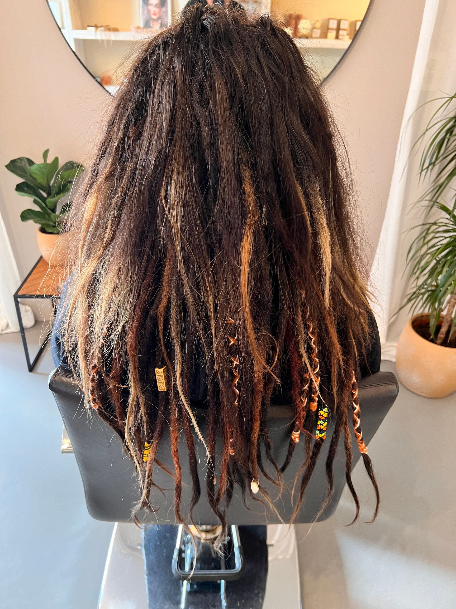 Een persoon zit op een stoel, met de rug naar de camera, en heeft lange, donkere dreadlocks met gouden accenten en kralen.