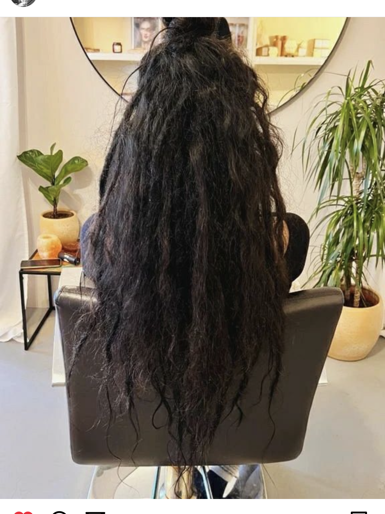 Persoon in een kappersstoel met lang, donker, gestructureerd haar; planten en een spiegel op de achtergrond.