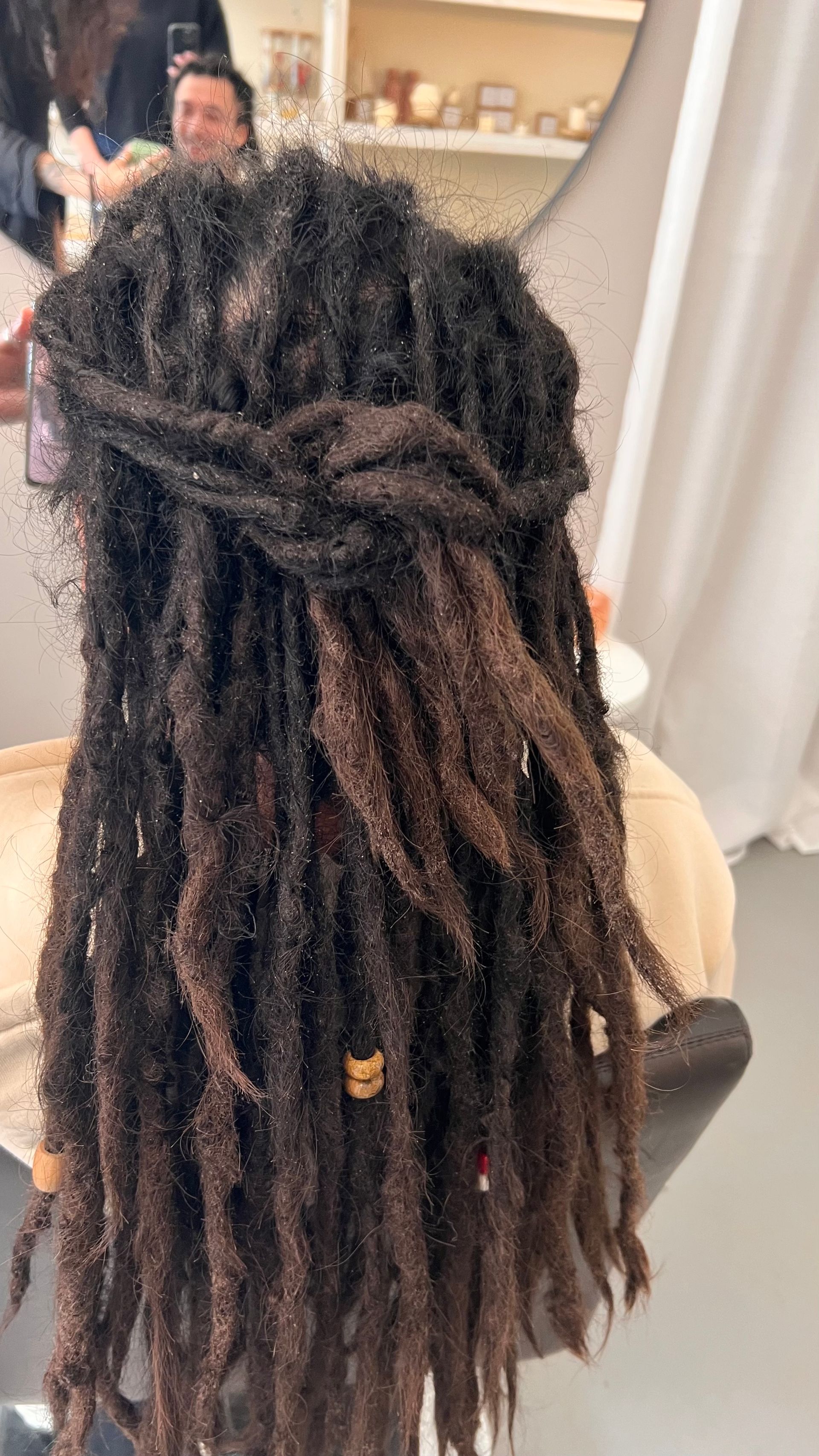 De persoon heeft dreadlocks die gedeeltelijk opgestoken zijn. Donkerbruin haar met houten kralen, zittend in een kapsalon.