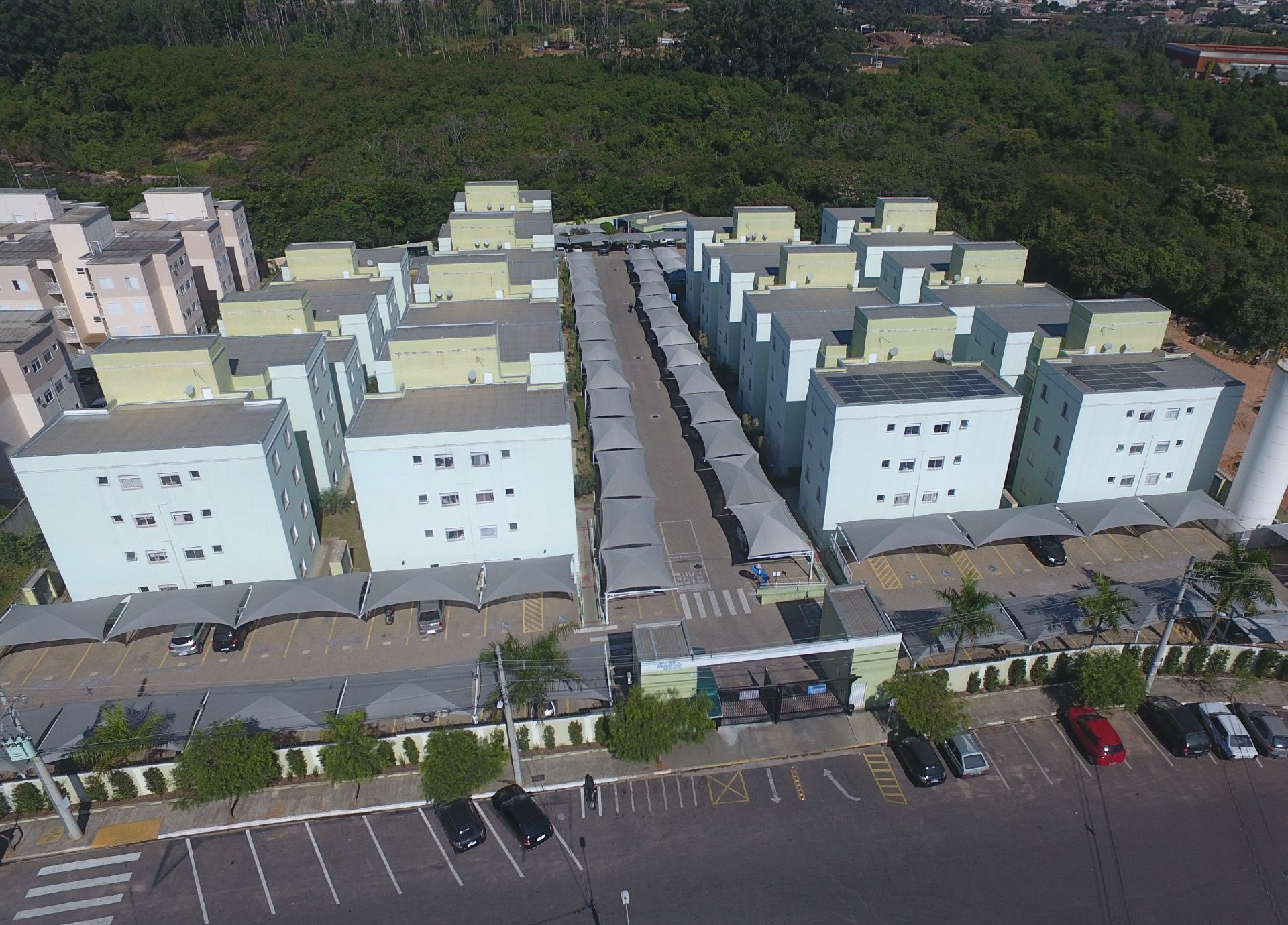 Vista aérea de um complexo habitacional com edifícios azuis claros, uma estrada central e uma floresta ao fundo.