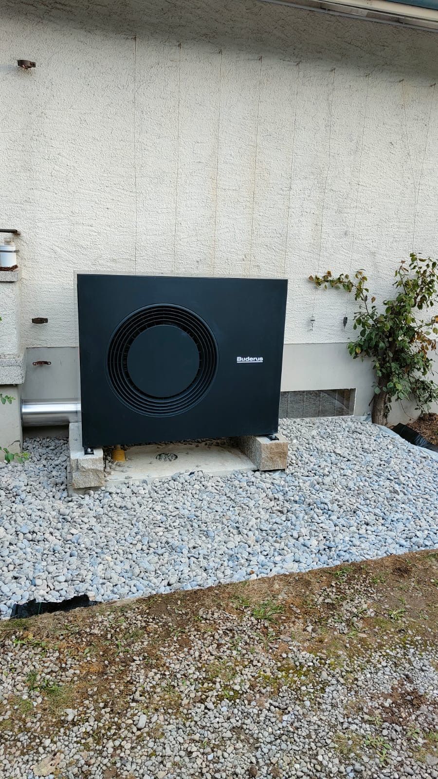 Schwarze Außenwärmepumpe auf Kies neben einer Hauswand, daneben ein kleiner Strauch