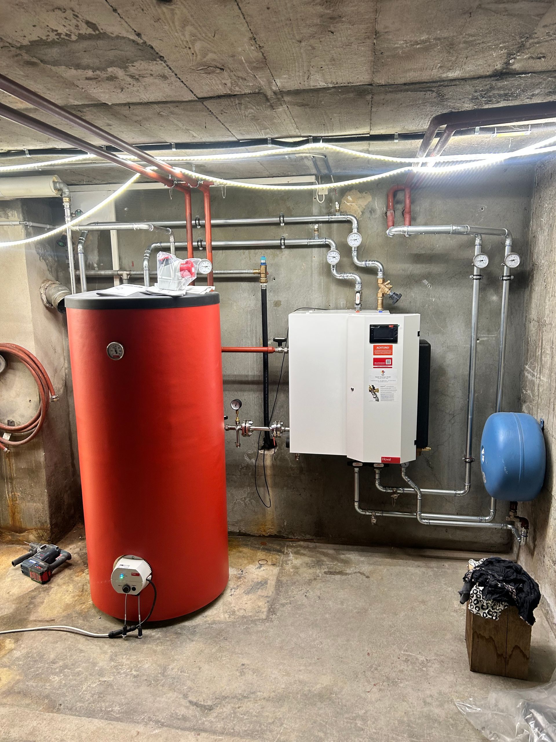 Hauswirtschaftsraum im Keller mit rotem Warmwasserbereiter, weißem Heizkessel, freiliegenden Rohren und blauem Ausdehnungsgefäß