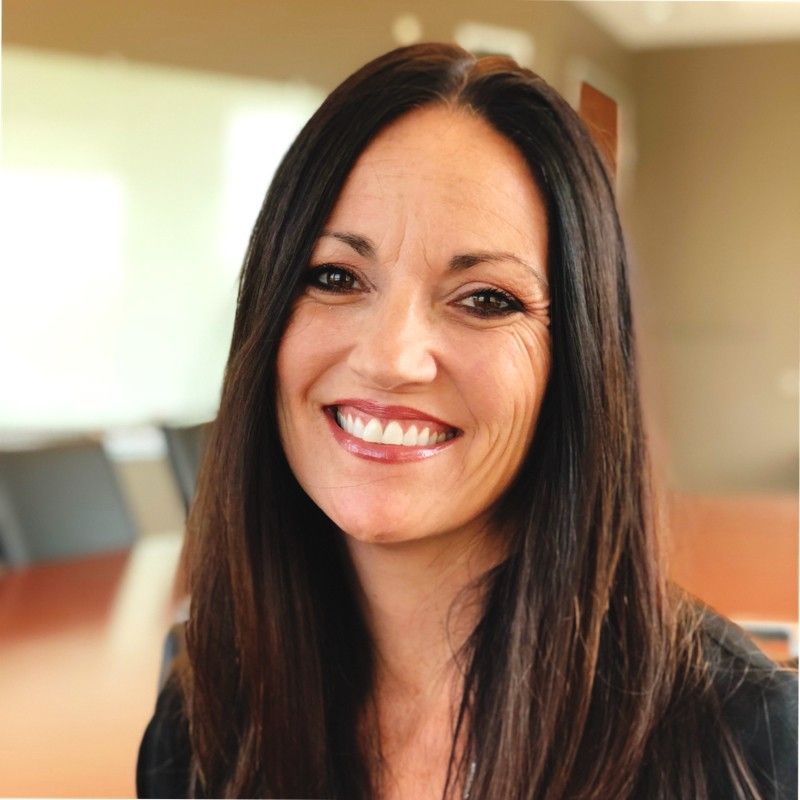 Smiling woman with long dark hair, wearing light makeup, photographed indoors with a softly blurred background.