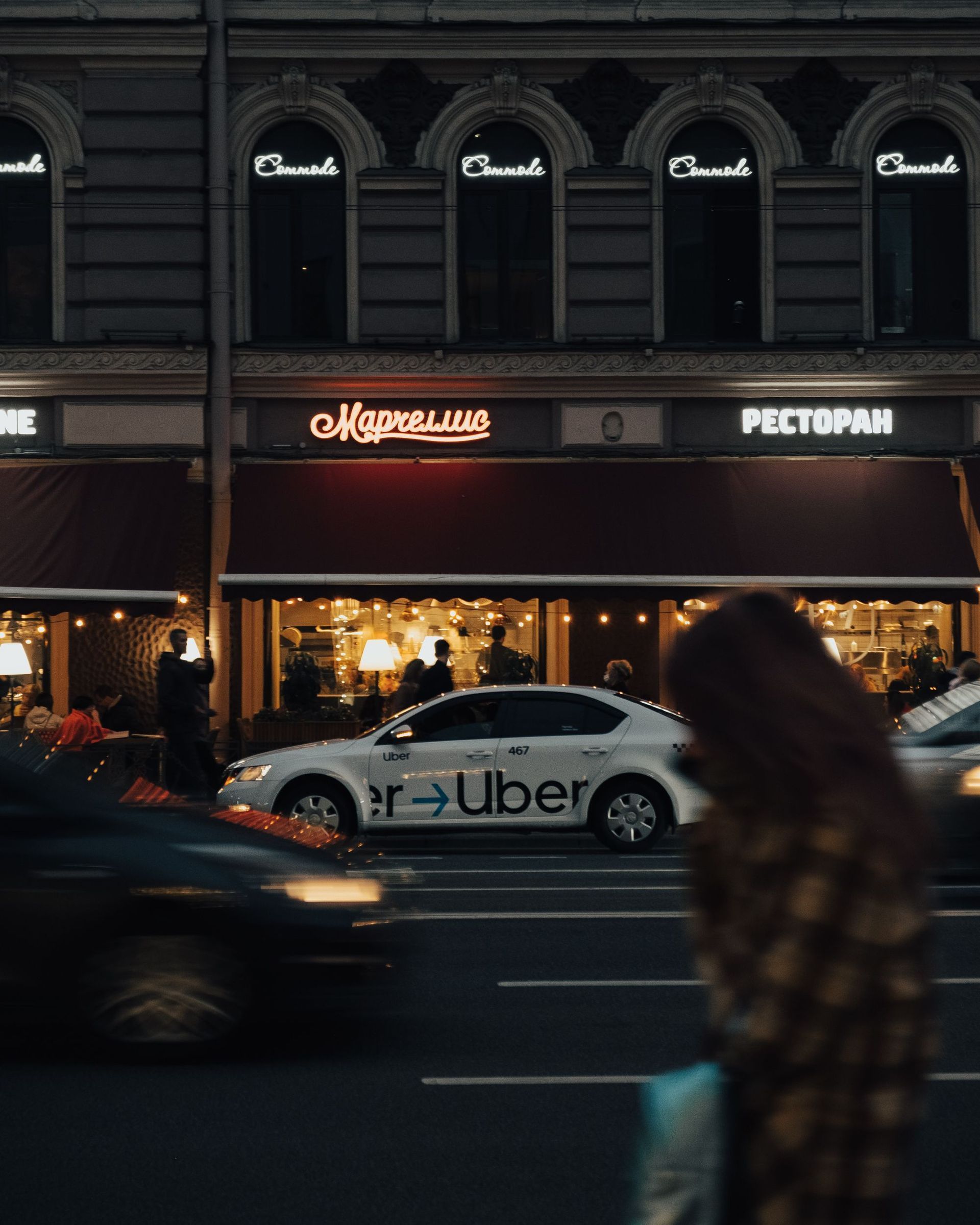 Nighttime city street with a lit restaurant storefront and an Uber car passing by