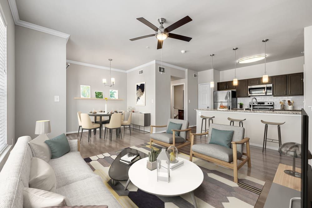 Spacious and modern open-plan living area with combined dining and kitchen space, featuring a light color palette. The lounge area includes a textured sofa with cushions, two wooden armchairs with teal cushions, a white round coffee table, and a patterned area rug. The dining area has a simple table set for six with a hanging chandelier above, while the contemporary kitchen boasts dark cabinets, stainless steel appliances, a kitchen island with bar stools, and pendant lighting. Decorative elements include wall art, plants, and minimalistic design touches throughout at Marquis at Stonebriar in Frisco, TX.