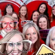 Group of smiling people posing with oversized glasses props in front of a red backdrop