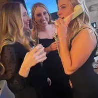 Three women in black dresses smiling and chatting at a party while holding drinks and one is talking on a rotary phone