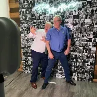 Two people posing in front of a black-and-white photo collage wall at an indoor event.