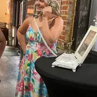 Woman in a colorful dress speaking on a vintage white phone beside a black table with a framed sign.