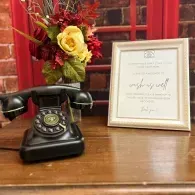 Black rotary phone and framed sign on a wooden desk with red floral arrangement behind them