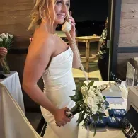 Bride in a white dress holding a bouquet and speaking on a phone at a reception table