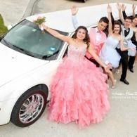 Young people in pink and white formalwear posing beside a white limousine, smiling and waving