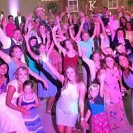 Crowd dancing at a pink-lit party, with many people raising their hands and smiling.