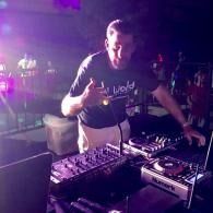 DJ at a mixing console under purple stage lights, giving a thumbs-up in a club setting