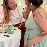 Two women chatting beside a table with a rotary phone and receiver at an event, one in a mint-green dress with tattoos and a name tag.