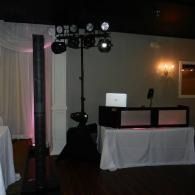 Dimly lit event room with DJ booth, laptop, lights, and draped tables