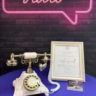 Vintage rotary phone, framed certificate, and business card on a purple table against a brick wall with neon sign.