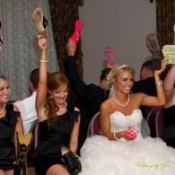 Women in dresses raise drinks and dance at a party, with one in a white bridal gown holding a pink drink, and each holding a single shoe in the air