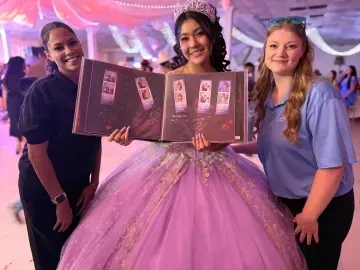 Three women pose with a glittery pink prom dress display at an indoor event.