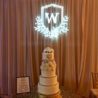 Wedding cake beneath glowing “W” sign and wreath on draped backdrop