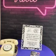 Vintage cream rotary phone on a purple table beside a framed sign under pink neon lighting.