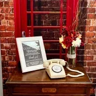 Vintage beige rotary phone on a wooden table beside a framed sign and red flowers against a brick wall.