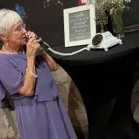 Woman in a purple dress speaking into the receiver of a vintage phone beside a white table display at an event
